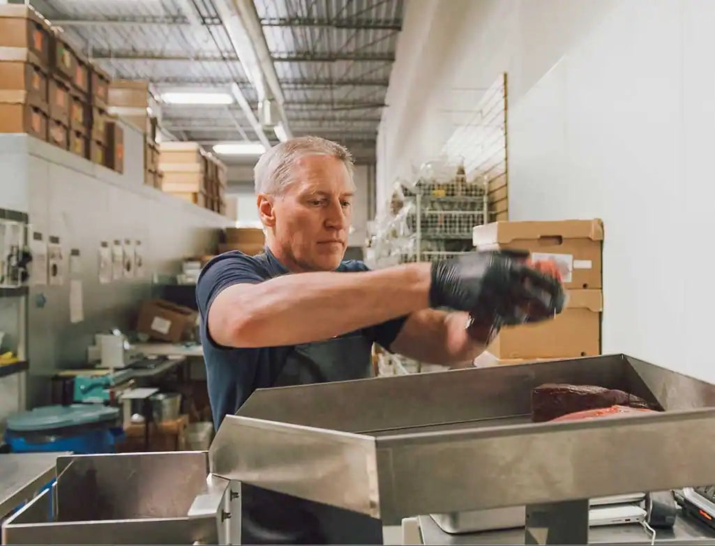 a man wearing gloves lifts raw meat out of a stainless steel tray