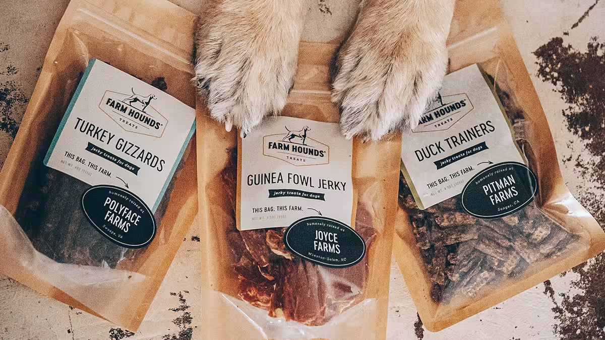 Three bags of Farm Hounds single ingredient dog treats arranged on a light surface, with a dog's paws resting on top.